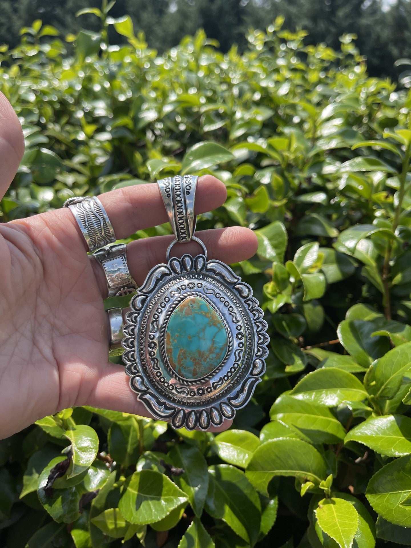 Native American Pilot Mountain Turquoise Pendant with intricate silver detailing held against green foliage background