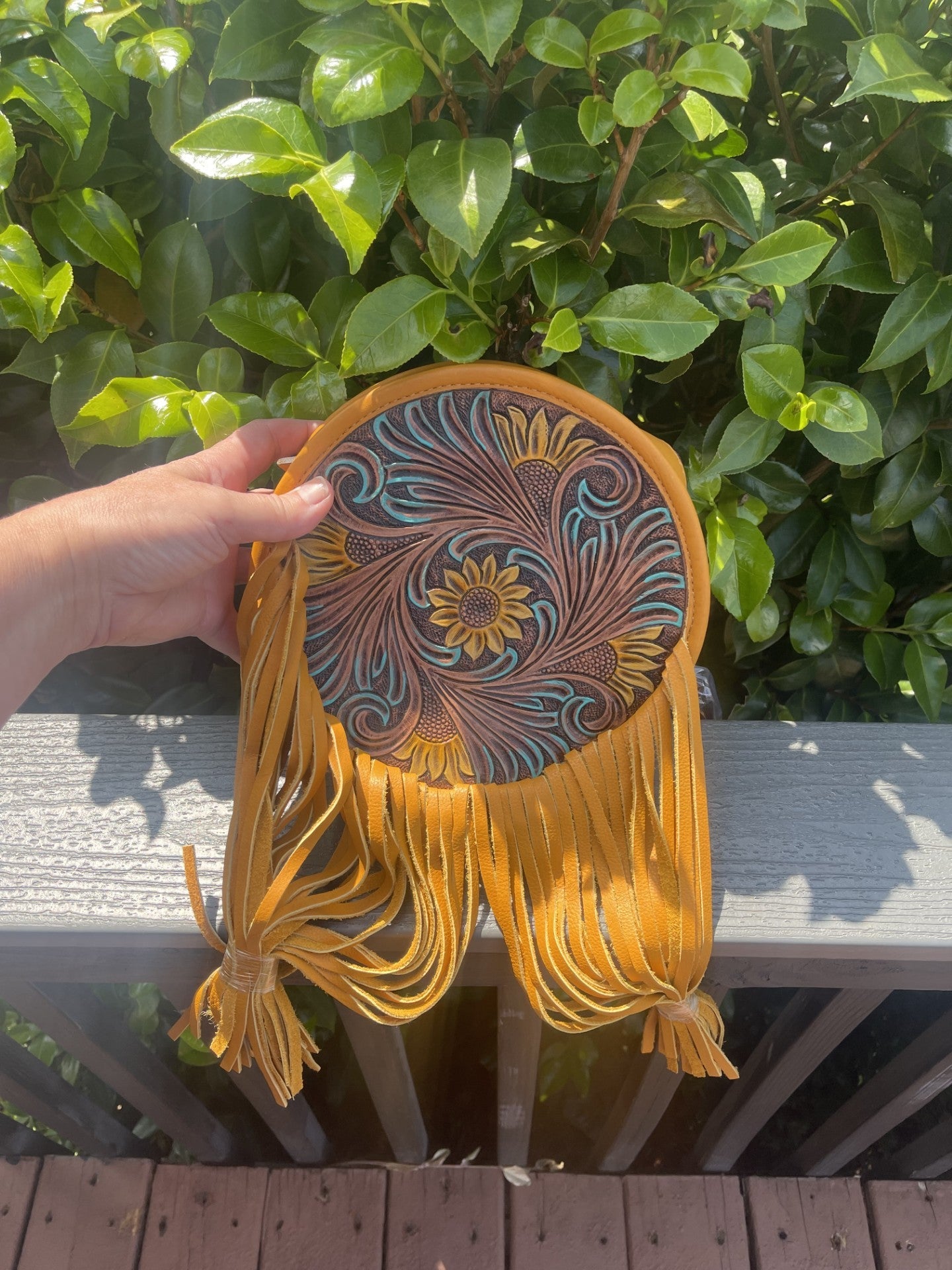 Yellowish-tan sunflower tooled canteen crossbody bag with fringe held outdoors against green foliage
