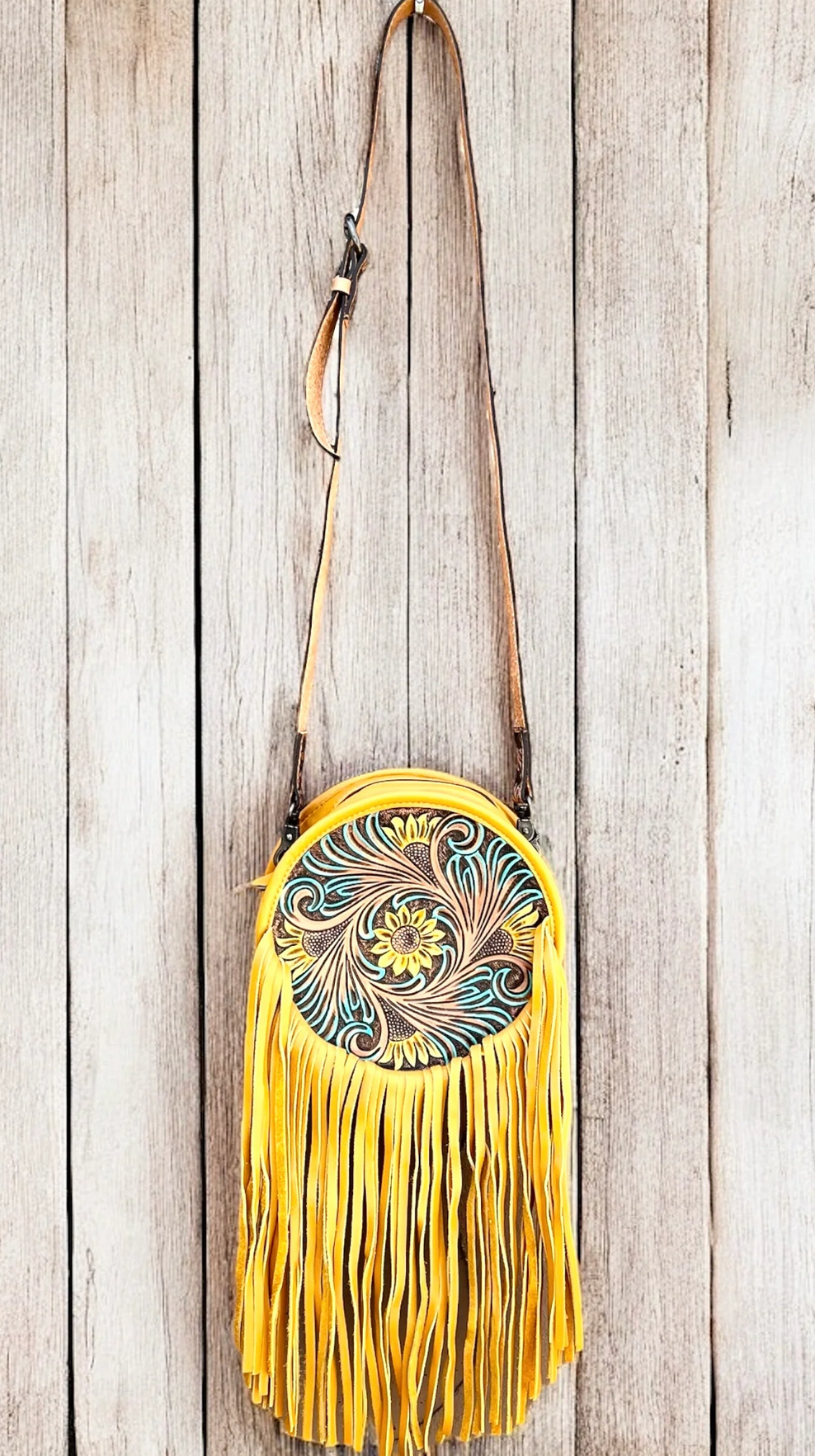 Yellowish-Tan sunflower tooled leather crossbody bag with fringe and canteen style strap against wooden background