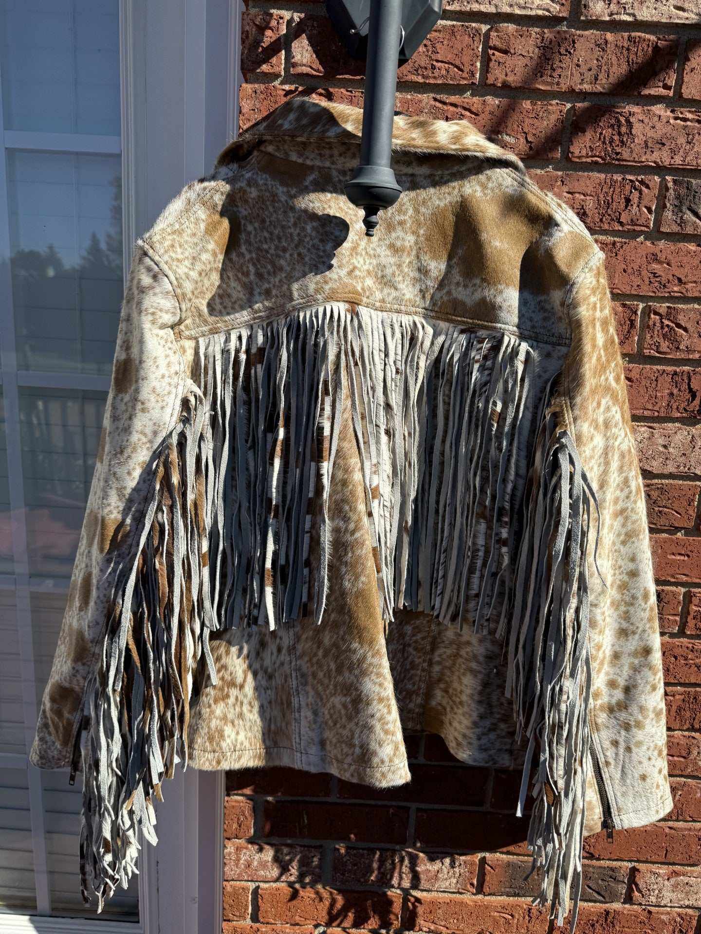 Tan cowhide jacket with fringe hanging on hanger against brick wall background