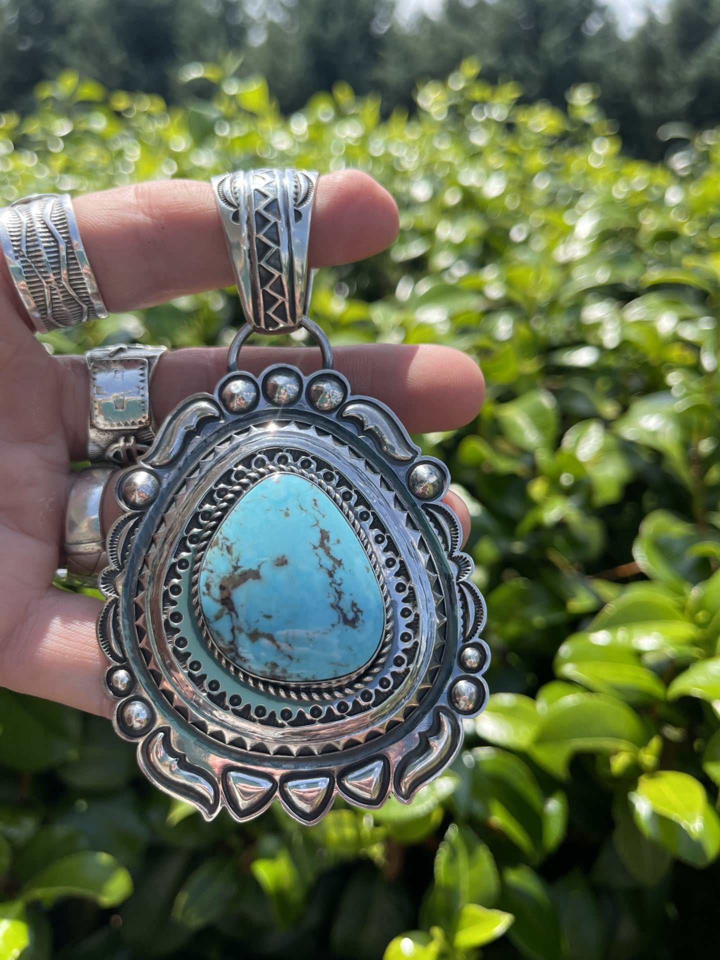 Native American Kingman Turquoise pendant with intricate silver detailing held against green foliage background