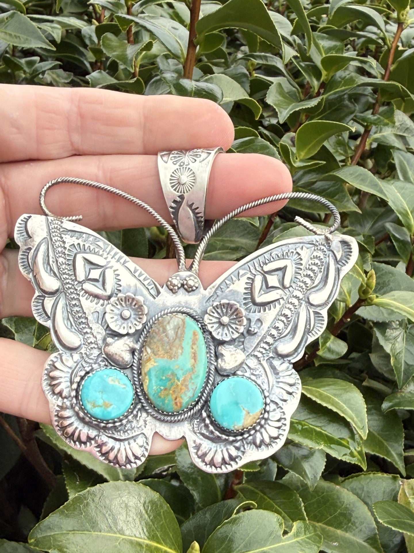 Kingman Butterfly Pendant by Richard Yazza silver butterfly pendant with turquoise stones and floral stamp work held against green leaves