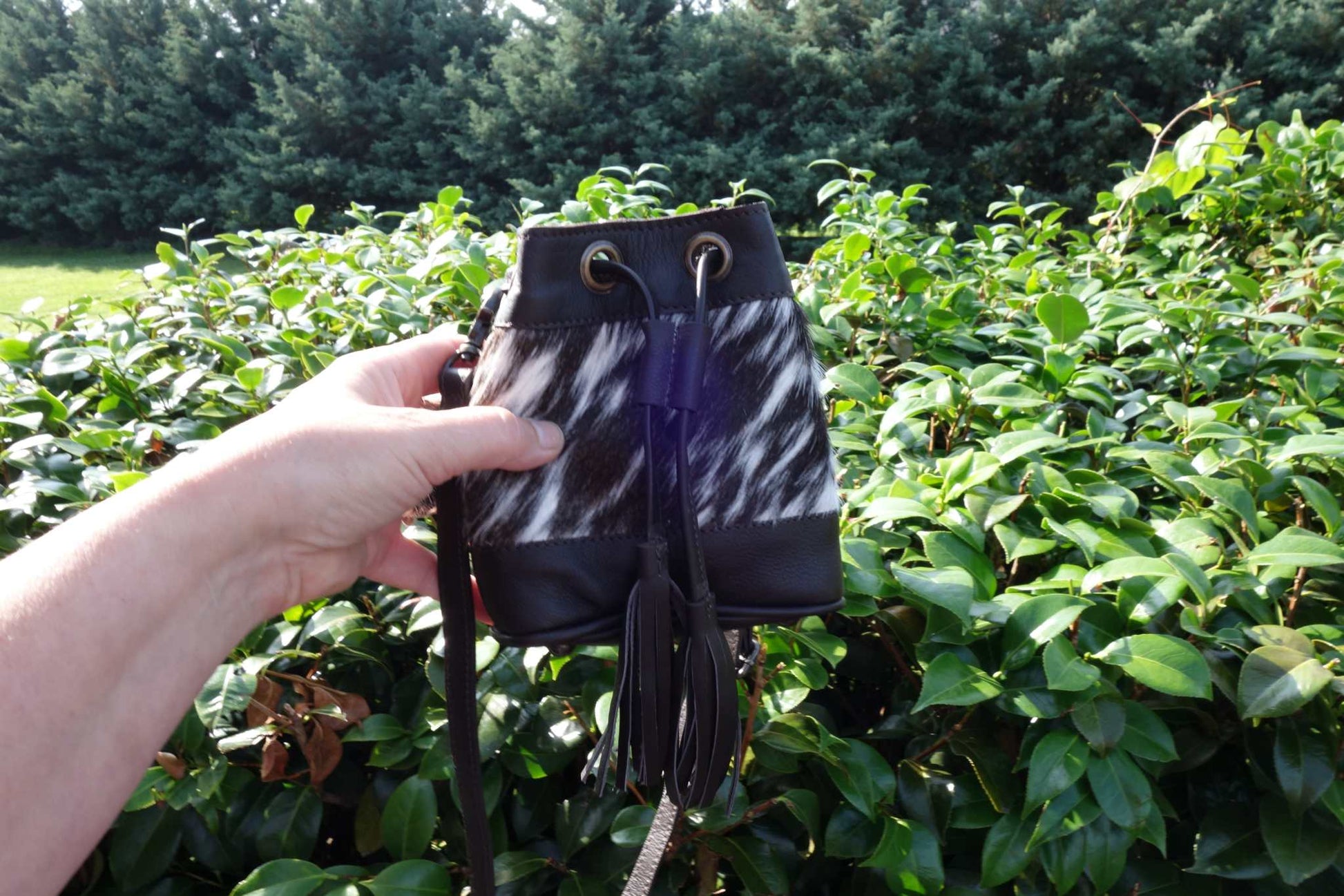 Hand holding a brown cowhide drawstring crossbody kids bag with black leather accents outdoors in front of green foliage