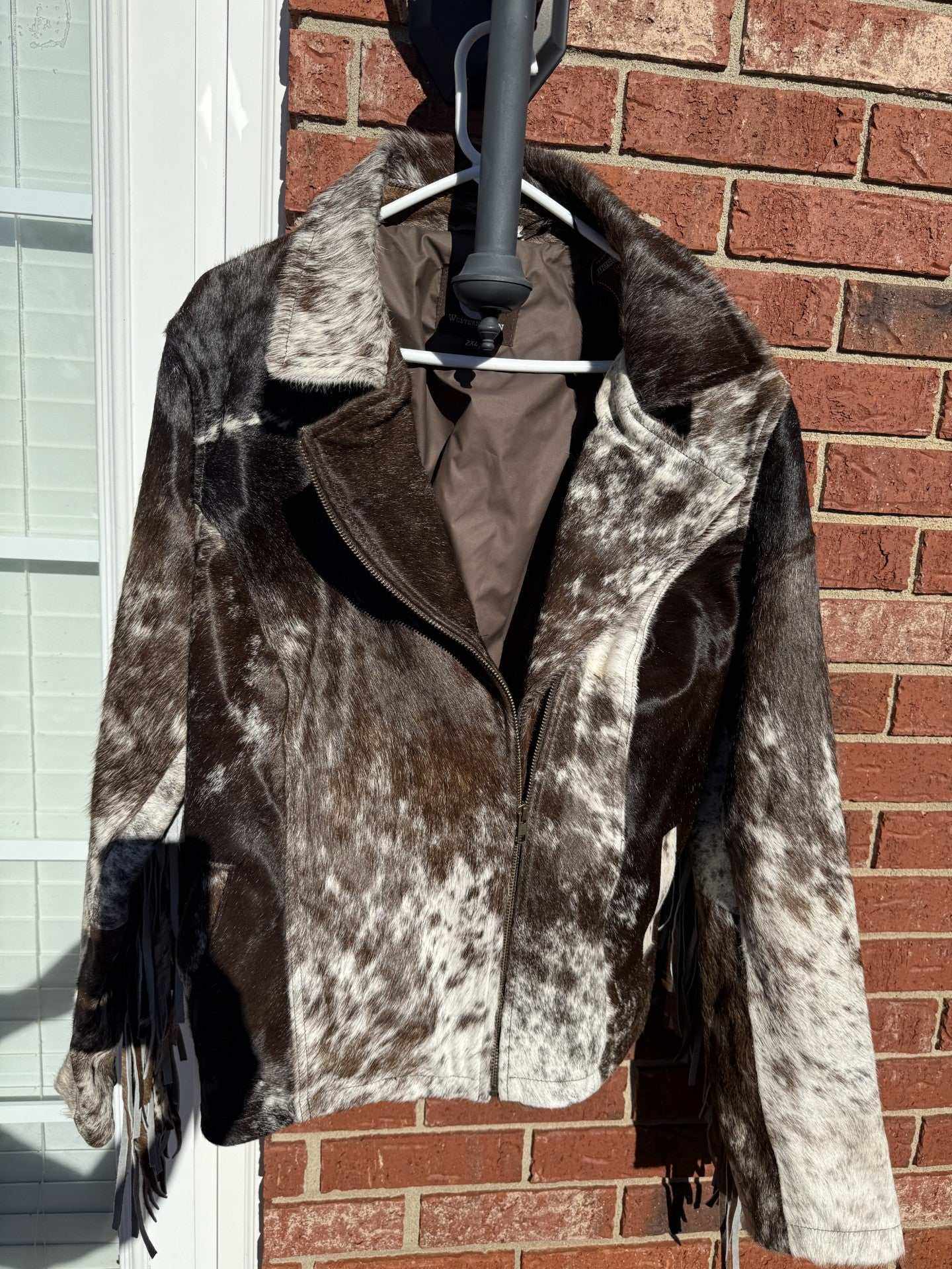 Brown cowhide leather jacket with fringe in 2XL size hanging outside on brick wall