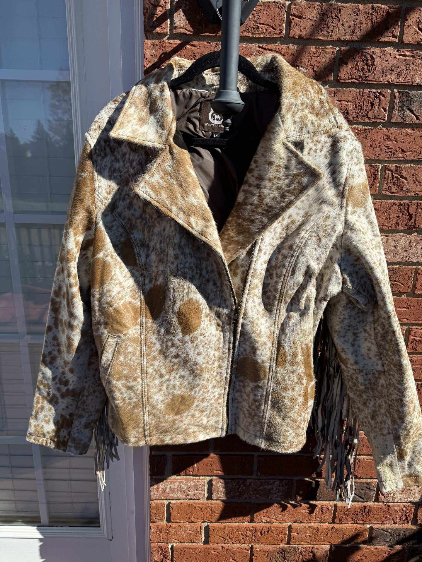 Tan cowhide jacket with fringe hanging on black hanger against brick wall background