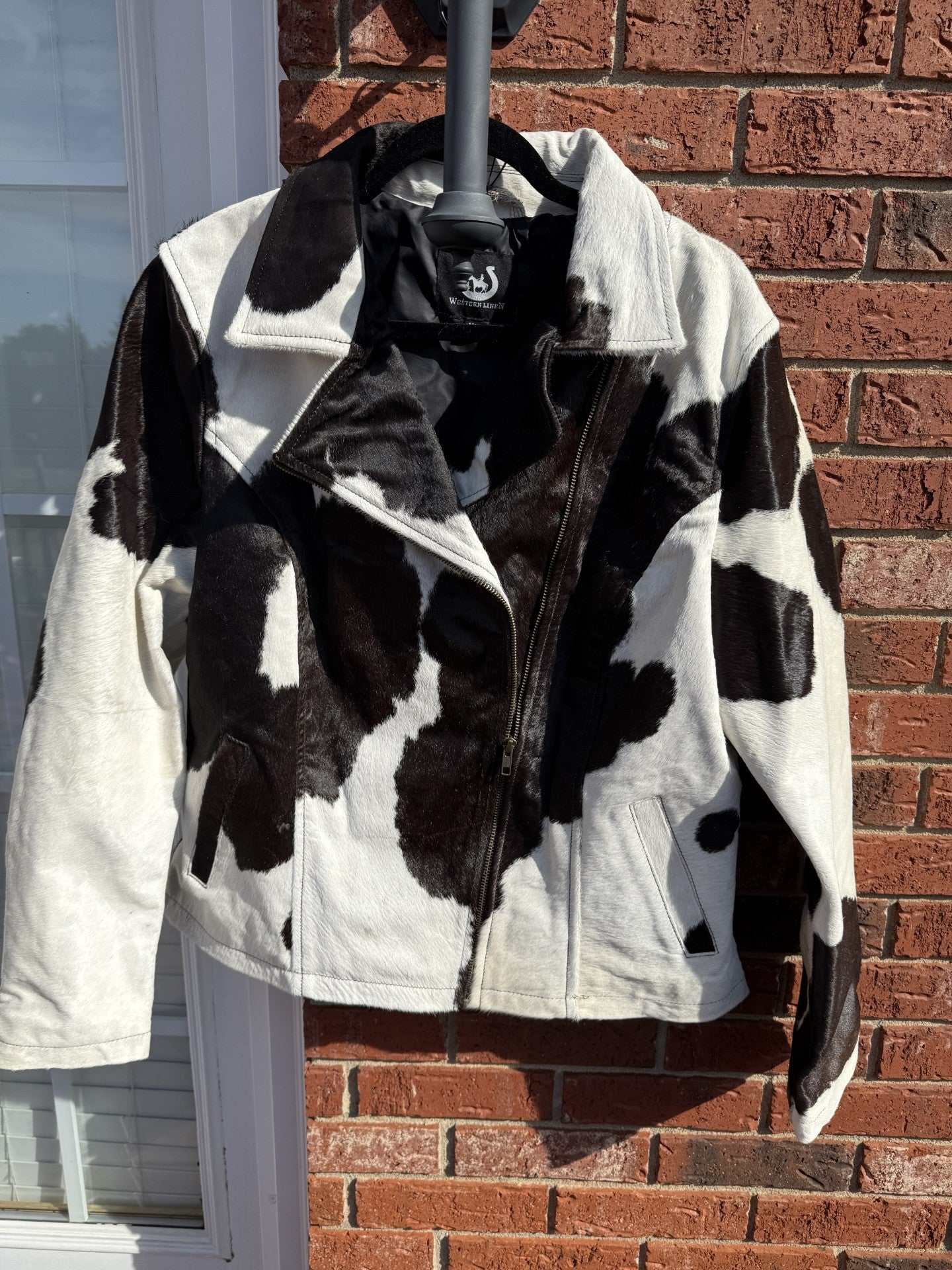 Black and white cowhide leather jacket XL with unique cow pattern and fringe details hanging against brick wall