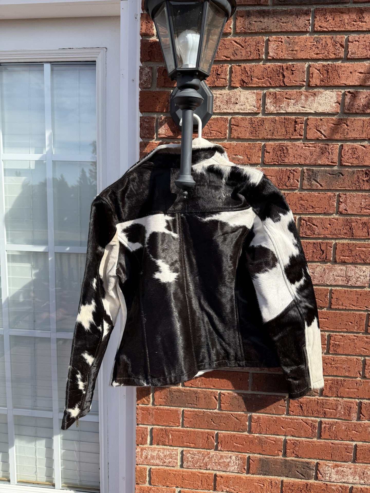 Black and white cowhide jacket hanging on outdoor lamp against brick wall, medium size women’s stylish jacket